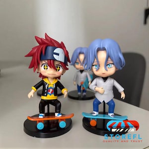 May include: Three anime-style figurines of characters on skateboards. The figurines have large eyes and colorful hair. One figure has red hair and a black jacket, another has blue hair and a white shirt. The skateboards are orange and blue.