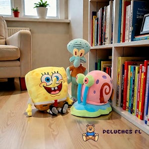 K&ouml;nnte beinhalten: Pl&uuml;schtiere mit Figuren aus der Zeichentrickserie SpongeBob Schwammkopf: SpongeBob, Thadd&auml;us und Gary die Schnecke. Die Spielzeuge befinden sich in einem Raum mit einem B&uuml;cherregal und einem Sofa.