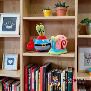 K&ouml;nnte beinhalten: Ein Holzregal zeigt Pl&uuml;schtiere von Mr. Krabs und Gary aus Spongebob Schwammkopf. Das Regal enth&auml;lt auch gerahmte Kunstwerke, Topfpflanzen und eine Sammlung von B&uuml;chern. Die Pl&uuml;schtiere sind farbenfroh und detailliert.