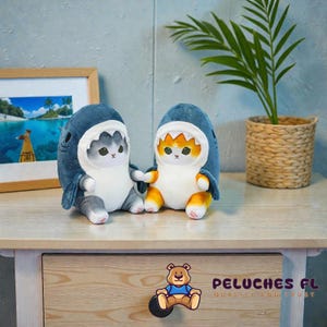 May include: Two plush cat toys wearing shark costumes. One cat is gray, the other is orange and white. The shark costumes are blue with white teeth. The toys are sitting on a wooden surface.