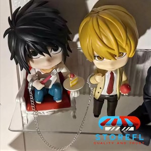 Puede incluir: Dos figuras de estilo anime, una con cabello negro sentada en una silla roja, sosteniendo un tenedor y un pastel, y la otra con cabello rubio sosteniendo una manzana. Ambos están encadenados.