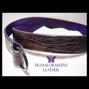 Puede incluir: Un cinturón de cuero marrón oscuro con una superficie texturizada y un interior morado. El cinturón tiene una hebilla plateada y el texto "TRANSFORMATIVE LEATHER" con un logotipo de mariposa.
