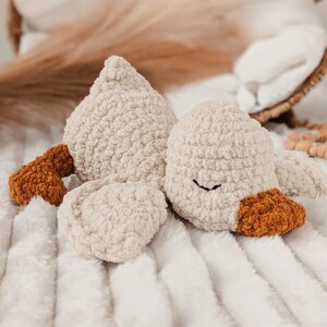 Puede incluir: Un peluche de ganso color crema con una apariencia texturizada y esponjosa. El ganso tiene detalles marrones en el pico y las patas, y un simple ojo cosido en negro. El juguete est&aacute; sobre una manta blanca texturizada.