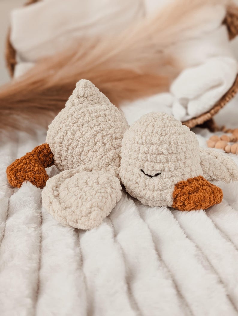Puede incluir: Un peluche de ganso color crema con una apariencia texturizada y esponjosa. El ganso tiene detalles marrones en el pico y las patas, y un simple ojo cosido en negro. El juguete est&aacute; sobre una manta blanca texturizada.
