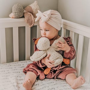 Puede incluir: Un beb&eacute; con un turbante y un mono color rubor est&aacute; sentado en una cuna blanca, agarrando un juguete de pato de ganchillo. El pato es de color crema con detalles en naranja. Otros juguetes de ganchillo est&aacute;n en la cabecera de la cuna.