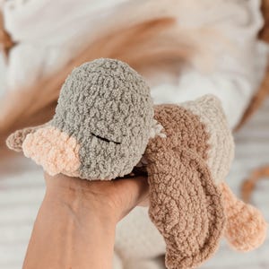 Puede incluir: Un juguete de pato tejido a mano en tonos grises, marr&oacute;n y melocot&oacute;n. El pato tiene una textura suave y ojos cerrados. El juguete se sostiene en una mano, mostrando su tacto suave y tierno. Un art&iacute;culo encantador y artesanal.