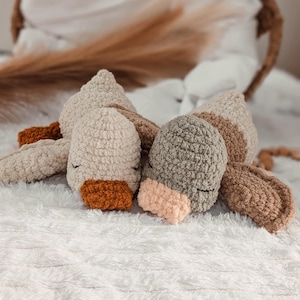 Puede incluir: Juguete de peluche tejido a ganchillo con forma de animal durmiente. El juguete presenta una cabeza beige claro, orejas marrones y un pico naranja. Est&aacute; sobre una superficie blanca y esponjosa.