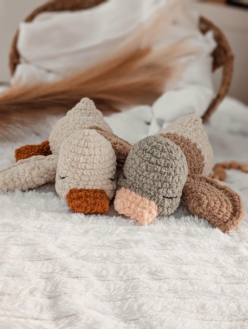 Puede incluir: Juguete de peluche tejido a ganchillo con forma de animal durmiente. El juguete presenta una cabeza beige claro, orejas marrones y un pico naranja. Est&aacute; sobre una superficie blanca y esponjosa.