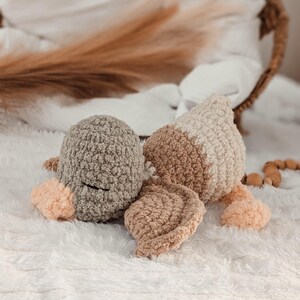 Puede incluir: Un juguete de pato de ganchillo hecho a mano en tonos grises, beige y melocot&oacute;n. El pato de peluche tiene un aspecto texturizado y esponjoso con cabeza, cuerpo y alas redondeadas. El juguete est&aacute; sobre una superficie blanca texturizada.