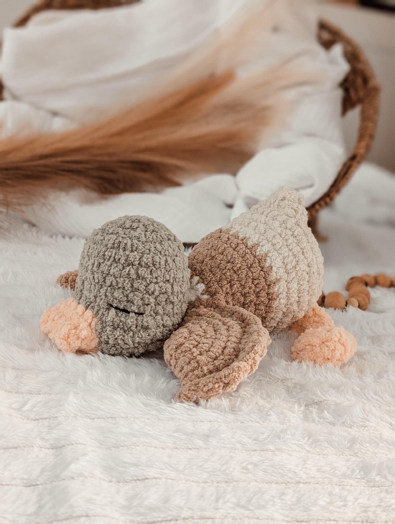 Puede incluir: Un juguete de pato de ganchillo hecho a mano en tonos grises, beige y melocot&oacute;n. El pato de peluche tiene un aspecto texturizado y esponjoso con cabeza, cuerpo y alas redondeadas. El juguete est&aacute; sobre una superficie blanca texturizada.