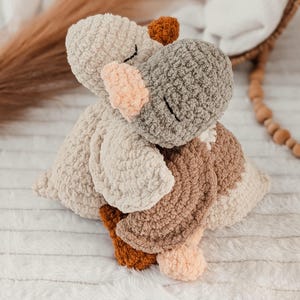 Puede incluir: Un juguete de hipop&oacute;tamo de peluche hecho a mano en tonos beige, gris, marr&oacute;n y melocot&oacute;n. El hipop&oacute;tamo est&aacute; tejido con un hilo suave y texturizado, con los ojos cerrados y un dise&ntilde;o suave y abrazable. El juguete est&aacute; sobre una superficie blanca y texturizada.