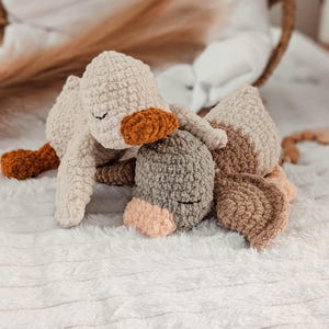 Puede incluir: Un juguete de ganso tejido a mano en crema, marr&oacute;n y gris. El ganso tiene un pico y patas naranjas, con los ojos cerrados, y est&aacute; sobre una superficie blanca texturizada. El juguete es suave y afelpado, ideal para una guarder&iacute;a o como objeto decorativo.