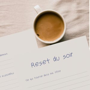 Può includere: Una tazza bianca piena di caffè è appoggiata su un foglio di carta bianco con il testo francese "Reset du soir" in scrittura blu. La carta ha linee per scrivere e altro testo francese.