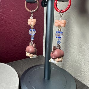 May include: Two keychains with a brown and white cartoon character charm. Each keychain has a different colored ring, one is red and the other is bronze. The charms are decorated with pink bows and blue and clear beads.