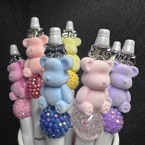 May include: A collection of decorative pens with colorful teddy bear toppers. The pens feature white barrels, sparkling jewel-encrusted bases, and small teddy bear figures in various pastel colors, including pink, blue, and purple. Each pen has a silver bow detail.
