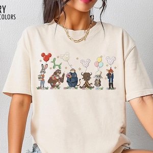 Puede incluir: Camiseta de color marfil con un gr&aacute;fico de dibujos animados de varios personajes de animales sosteniendo globos. Los globos son de varias formas y colores. El texto "IVORY COMFORT COLORS" est&aacute; impreso en la parte superior izquierda.