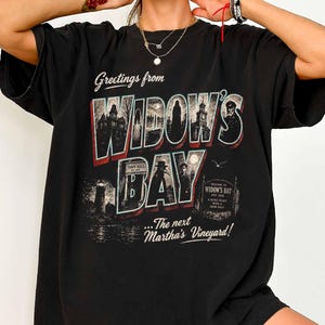Könnte beinhalten: Ein schwarzes Kurzarm-T-Shirt mit einem Grafikdruck und dem Text Greetings from WIDOW'S BAY ...The next Martha's Vineyard! Das Design zeigt Illustrationen eines Rathauses, eines Leuchtturms und Figuren in einer dunklen, stimmungsvollen Umgebung.