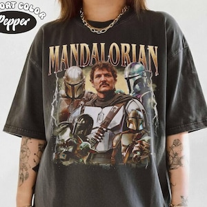 K&ouml;nnte beinhalten: Dunkelgraues T-Shirt mit einer Grafik des Mandalorianers und dem goldenen Text "MANDALORIAN". Das Shirt hat einen Vintage-Look, mit dem Comfort Color und Pepper Logo in der oberen linken Ecke.