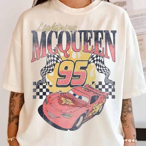 May include: Ivory-colored t-shirt featuring a graphic of Lightning McQueen from the movie Cars. The design includes the text "Lightning McQueen 95" with checkered flags and a red car illustration. The shirt is labeled "Comfort Colors".