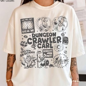 Può includere: T-shirt color avorio con un design grafico con il testo "Dungeon Crawler Carl" e varie illustrazioni. La maglietta include anche le frasi "You Will Not Break Me", "This Is An Outrage" e "Goddamnit Donut!"
