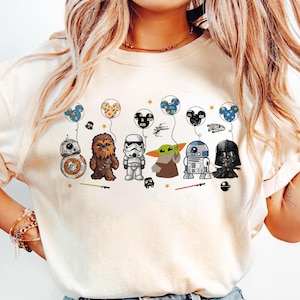 Peut inclure: T-shirt cr&egrave;me avec des personnages de Star Wars : BB-8, Chewbacca, un Stormtrooper, Grogu, R2-D2 et Dark Vador. Chaque personnage tient un ballon avec une t&ecirc;te de Mickey. Le t-shirt a une coupe d&eacute;contract&eacute;e.