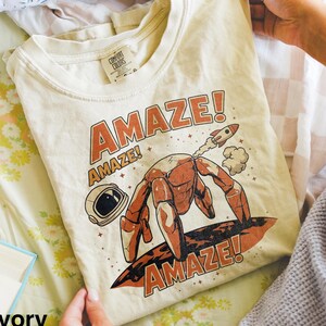 May include: A cream-colored t-shirt featuring a graphic print of a robotic creature with multiple legs standing on a planet surface, a small rocket, and an astronaut helmet. The text "AMAZE!" is printed in large orange letters above and below.