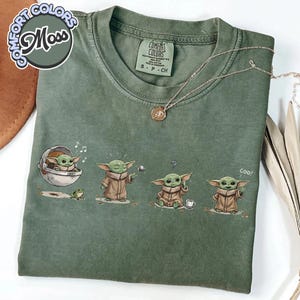 K&ouml;nnte beinhalten: Moosgr&uuml;nes T-Shirt mit einer Cartoonfigur in verschiedenen Posen. Die Figur wird mit einem Frosch, einem Ball und einer Tasse gezeigt. Auf dem T-Shirt stehen die W&ouml;rter "Comfort Colors" und "Moss".