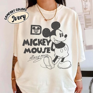 May include: Ivory-colored t-shirt featuring a classic Mickey Mouse graphic. The design includes the words "Mickey Mouse" in a bold, vintage font, with Mickey Mouse himself in a black and white illustration. The shirt also has the words "Comfort Colors" and "Ivory" printed on it.