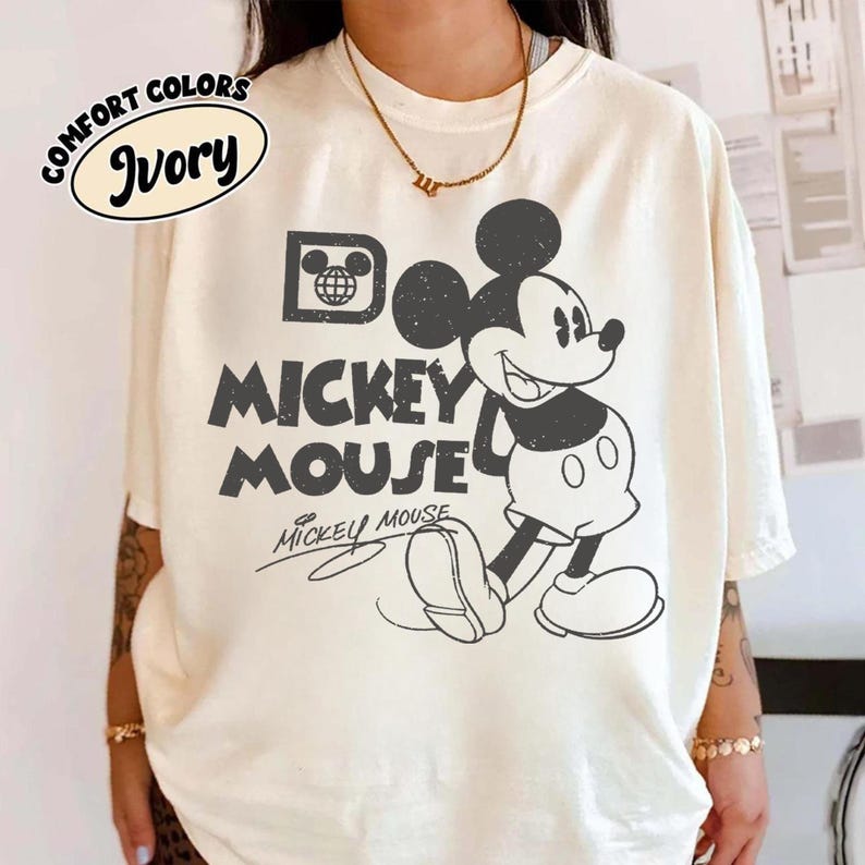May include: Ivory-colored t-shirt featuring a classic Mickey Mouse graphic. The design includes the words "Mickey Mouse" in a bold, vintage font, with Mickey Mouse himself in a black and white illustration. The shirt also has the words "Comfort Colors" and "Ivory" printed on it.