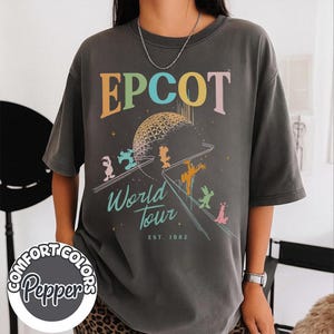 May include: Dark gray t-shirt with the word "EPCOT" in colorful letters above a graphic of a geodesic sphere and cartoon characters. The text "World Tour" and "EST. 1982" are below the graphic. The shirt has a Comfort Colors Pepper logo.