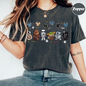 Peut inclure: T-shirt gris fonc&eacute; avec des personnages de dessins anim&eacute;s Star Wars tenant des ballons. Les personnages incluent BB-8, Chewbacca, un Stormtrooper, Grogu, R2-D2 et Dark Vador. Les ballons ont divers motifs, dont des oreilles de Mickey Mouse et des plan&egrave;tes.
