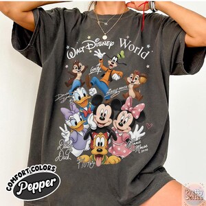 May include: Dark grey t-shirt featuring a graphic of Disney characters including Mickey Mouse, Minnie Mouse, Donald Duck, and Goofy. The shirt has the text "Walt Disney World" and character signatures. The shirt is labeled "Comfort Colors Pepper".