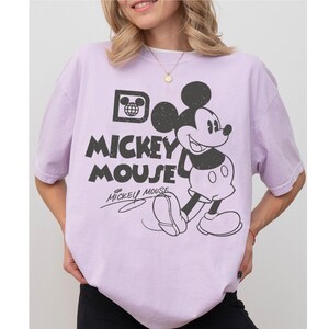 May include: A light purple t-shirt featuring a black and white graphic of Mickey Mouse. The design includes the words "Mickey Mouse" in a distressed font, along with Mickey's signature. The shirt has short sleeves and a relaxed fit.