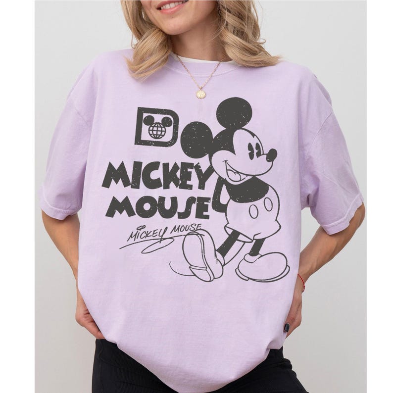May include: A light purple t-shirt featuring a black and white graphic of Mickey Mouse. The design includes the words "Mickey Mouse" in a distressed font, along with Mickey's signature. The shirt has short sleeves and a relaxed fit.