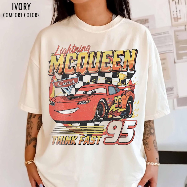 Peut inclure: T-shirt ivoire avec un motif de Flash McQueen du film Cars. Le design comprend le texte "Lightning McQueen", "Cars", "95" et "Think Fast". Le t-shirt est de style confort.