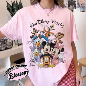 May include: A light pink t-shirt featuring a graphic of Disney characters, including Mickey Mouse, Minnie Mouse, Donald Duck, and Goofy. The words "Walt Disney World" are above the characters. The shirt also has the words "Comfort Colors" and "Blossom" printed on it.