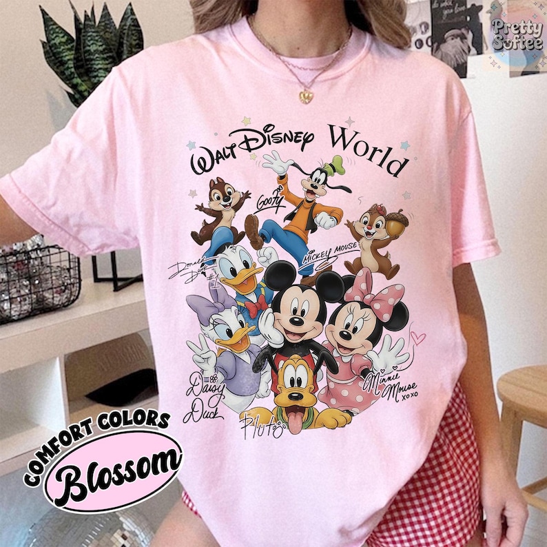 May include: A light pink t-shirt featuring a graphic of Disney characters, including Mickey Mouse, Minnie Mouse, Donald Duck, and Goofy. The words "Walt Disney World" are above the characters. The shirt also has the words "Comfort Colors" and "Blossom" printed on it.