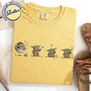 K&ouml;nnte beinhalten: Senfgelbes T-Shirt mit einer Grafik eines gr&uuml;nen Alien-Charakters in verschiedenen Posen. Auf dem Shirt stehen die W&ouml;rter "Comfort Colors" und "Butter". Das Shirt liegt neben einem Paar brauner Sandalen.