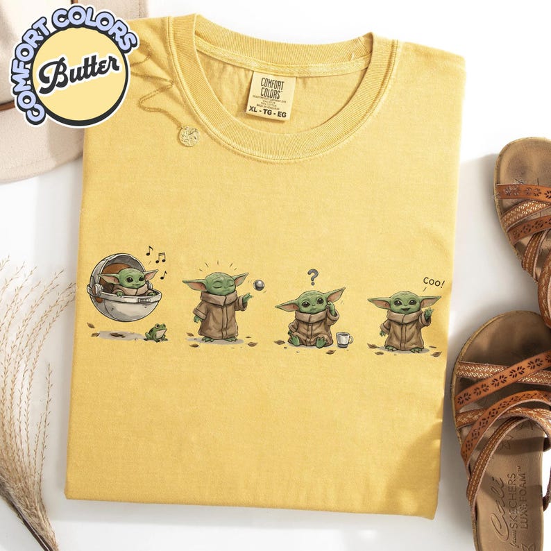 K&ouml;nnte beinhalten: Senfgelbes T-Shirt mit einer Grafik eines gr&uuml;nen Alien-Charakters in verschiedenen Posen. Auf dem Shirt stehen die W&ouml;rter "Comfort Colors" und "Butter". Das Shirt liegt neben einem Paar brauner Sandalen.