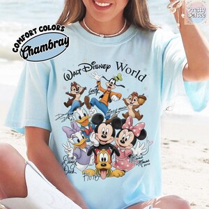 May include: Light blue t-shirt featuring a graphic of Disney characters including Mickey Mouse, Minnie Mouse, Donald Duck, and Goofy. The shirt has the words "Walt Disney World" and "Comfort Colors Chambray" printed on it.