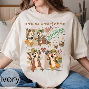May include: Ivory-colored t-shirt featuring a Disney Animal Kingdom graphic. The design includes cartoon chipmunks in safari attire, holding balloons and maps. The shirt has a crew neck and short sleeves.