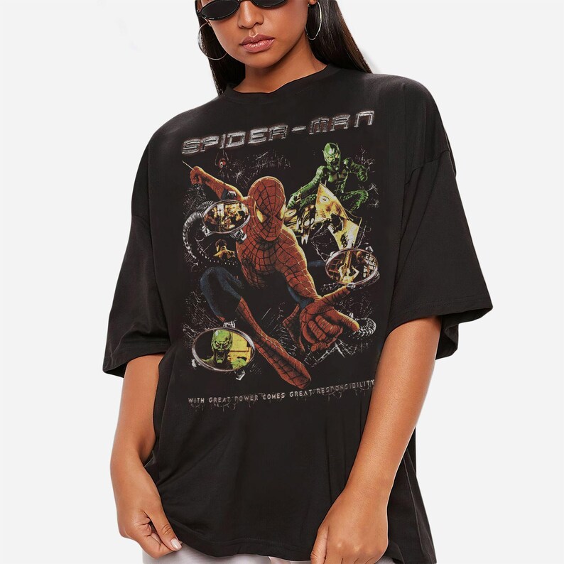 May include: Black t-shirt featuring a Spider-Man graphic. The design includes the words "SPIDER-MAN" at the top and the phrase "WITH GREAT POWER COMES GREAT RESPONSIBILITY" at the bottom. The graphic depicts Spider-Man and the Green Goblin.