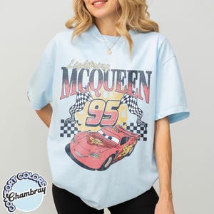 May include: Light blue t-shirt featuring a graphic of Lightning McQueen race car with the number 95. The design includes the text "Lightning McQueen" and checkered flags. The shirt is labeled "Comfort Colors Chambray".