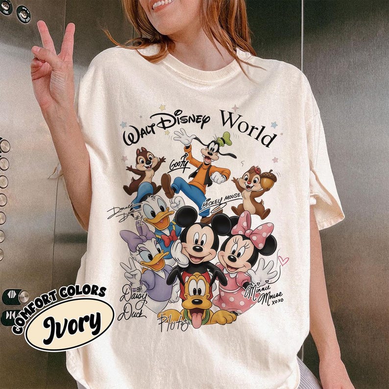 May include: Ivory-colored t-shirt featuring a graphic of Disney characters including Mickey Mouse, Minnie Mouse, Donald Duck, and Goofy. The text "Walt Disney World" is above the characters. The shirt also has the text "Comfort Colors" and "Ivory".