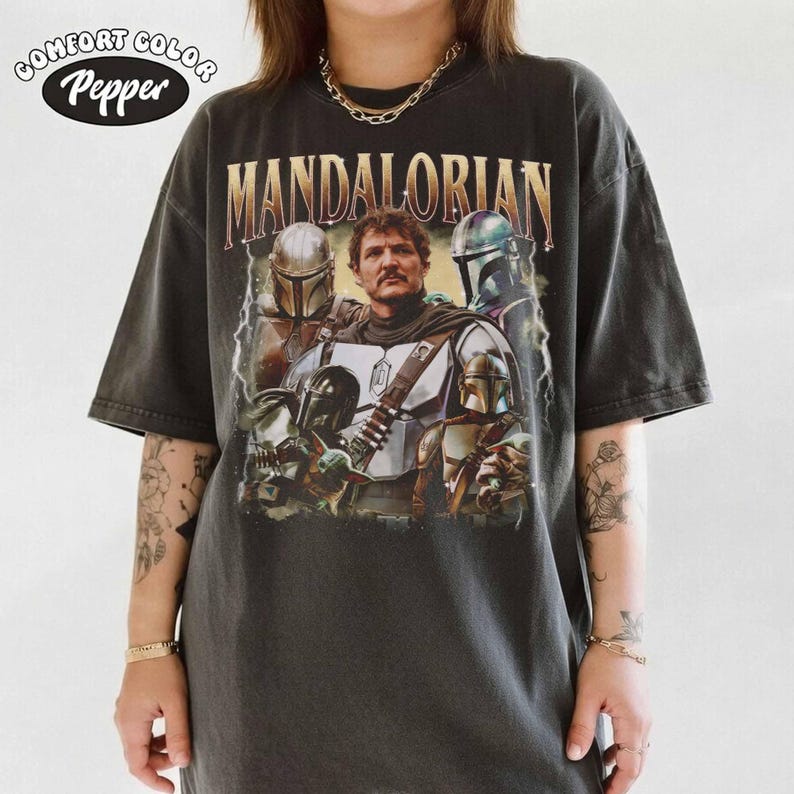 K&ouml;nnte beinhalten: Dunkelgraues T-Shirt mit einer Grafik des Mandalorianers und dem goldenen Text "MANDALORIAN". Das Shirt hat einen Vintage-Look, mit dem Comfort Color und Pepper Logo in der oberen linken Ecke.