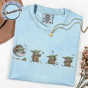 K&ouml;nnte beinhalten: Hellblaues T-Shirt mit einer Comicfigur in verschiedenen Posen, mit einem kleinen Frosch und einer Tasse. Eine goldene Sternkette liegt auf dem T-Shirt. Das T-Shirt hat das Etikett "Comfort Colors".