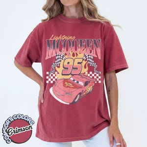 May include: Crimson red t-shirt featuring a graphic of Lightning McQueen from the Cars movie. The design includes the text "Lightning McQueen 95" with checkered flags and a cartoon image of the race car. The shirt has a vintage, worn look.