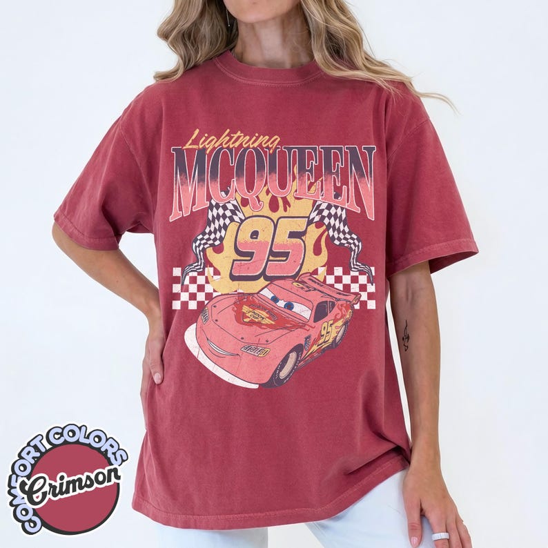 May include: Crimson red t-shirt featuring a graphic of Lightning McQueen from the Cars movie. The design includes the text "Lightning McQueen 95" with checkered flags and a cartoon image of the race car. The shirt has a vintage, worn look.
