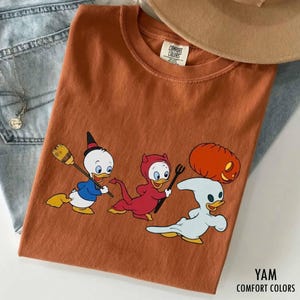 以下が含まれることがあります： ハロウィンの仮装をした3羽の漫画のキャラクターのアヒルが描かれた、錆色のTシャツ。1羽はほうきを持った魔女、1羽は熊手を持った赤い悪魔、もう1羽はカボチャを運ぶ幽霊の姿です。TシャツにはComfort Colorsのラベルが付いています。