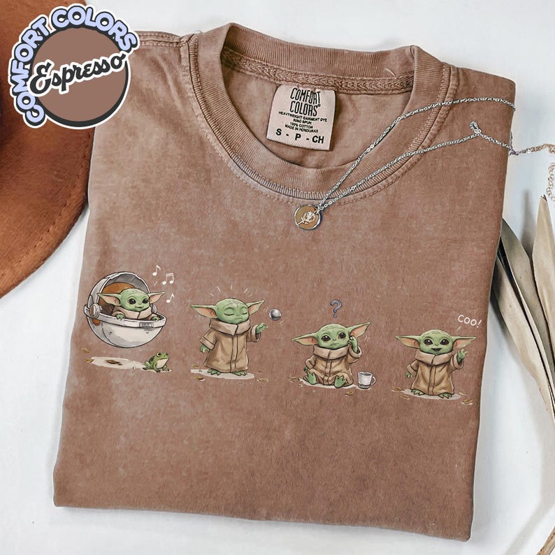 K&ouml;nnte beinhalten: Espressofarbenes T-Shirt mit Rundhalsausschnitt und kurzen &Auml;rmeln. Das Shirt zeigt eine Grafik eines gr&uuml;nen Alien-Charakters in verschiedenen Posen. Eine silberne Halskette mit einem kleinen Anh&auml;nger befindet sich auf dem Shirt. Die Marke "Comfort Colors" steht auf dem Shirt.